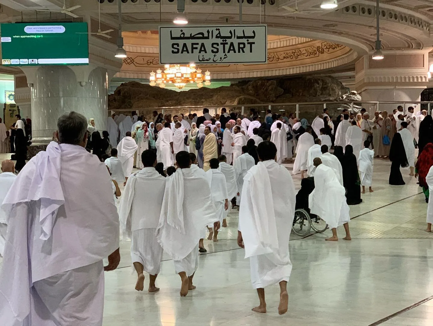 The interior of the sa'i gallery at the Masjid al-Haram, a wide marble hall under a vaulted ceiling with ceiling fans and a chandelier, pilgrims in white ihram walking towards the exposed rock of the hill of Safa behind a glass barrier, and a hanging sign reading Safa Start in Arabic and English