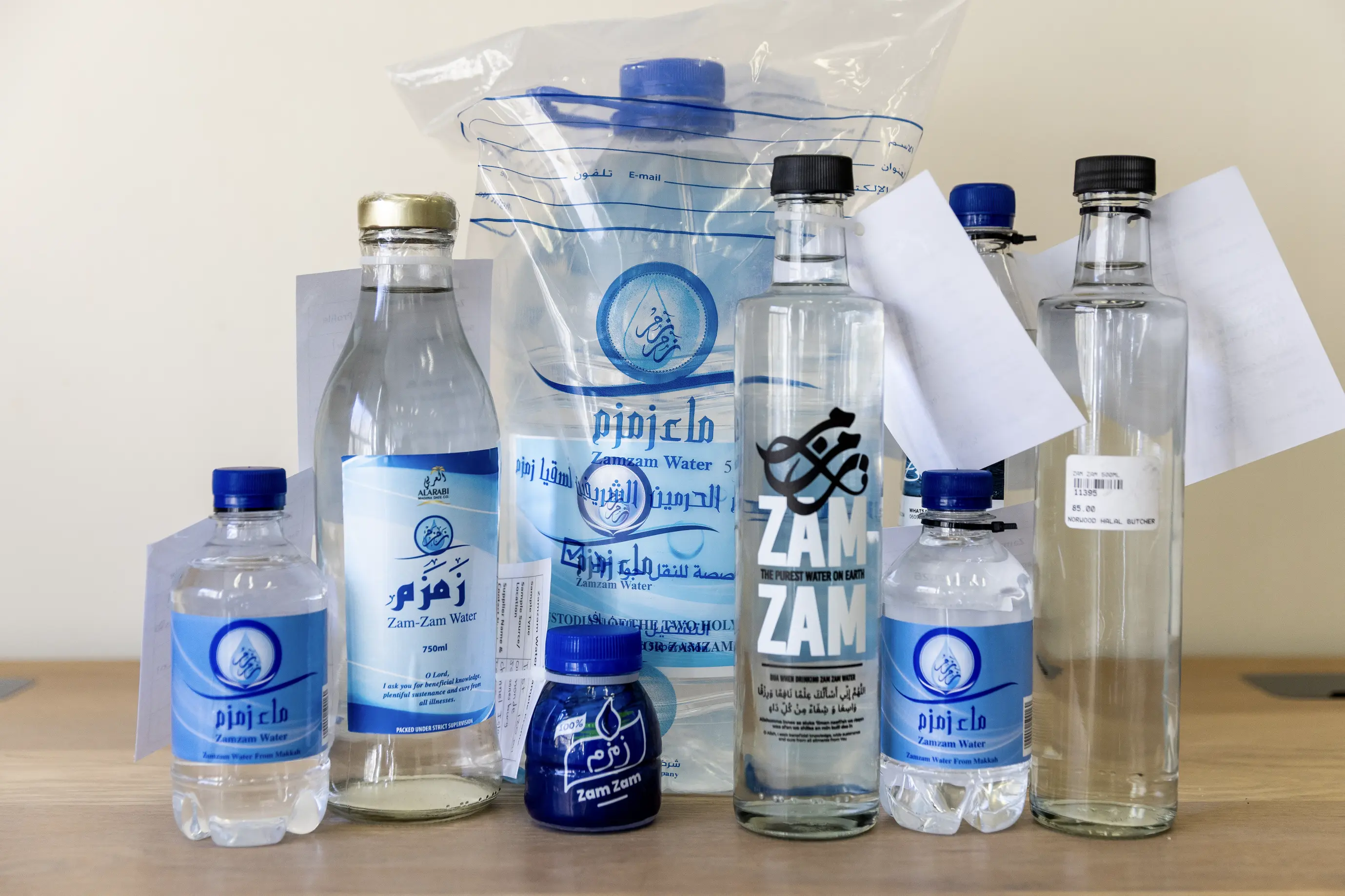 A group of clear bottles and a plastic pouch of drinking water arranged on a wooden surface, several labelled in Arabic and English as Zamzam water, with sample tags attached, against a plain pale wall